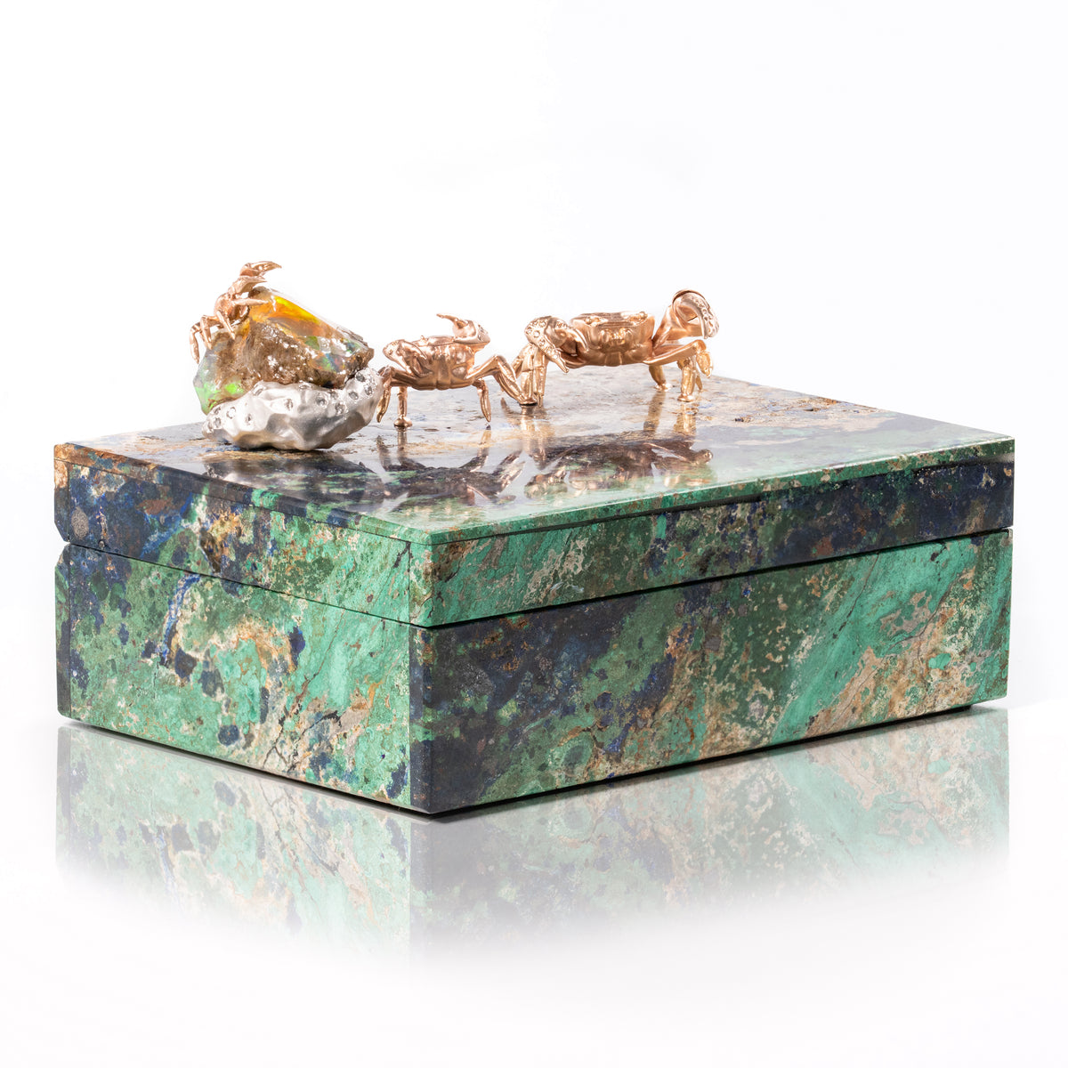 A rectangular jewelry box made of green and black marbled stone with three small gold crab figurines on top, photographed on a clean white background with a soft reflection below.