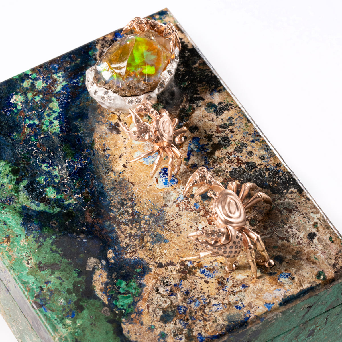 A close-up of jewelry displayed on a polished stone slab: two rose-gold crab-shaped earrings and a ring with an iridescent green-yellow stone, set against a white background.