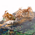 Three metallic crab figurines in copper and silver tones arranged on a marbled green and black stone slab against a white background.